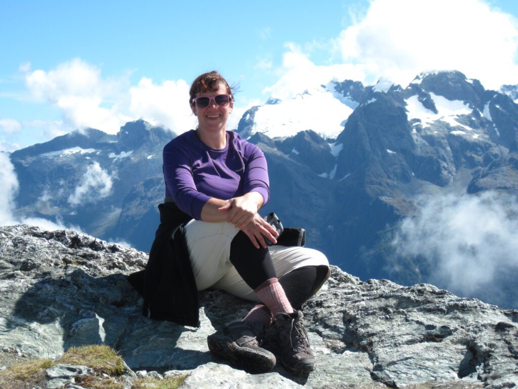 Anna Rankin sits on a mountaintop wearing a pair of pink sunglasses with the mountains behind her.
