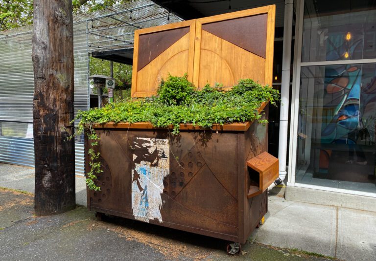 A brown dumpster sits in an alley with green plants growing out of it and over the edge.