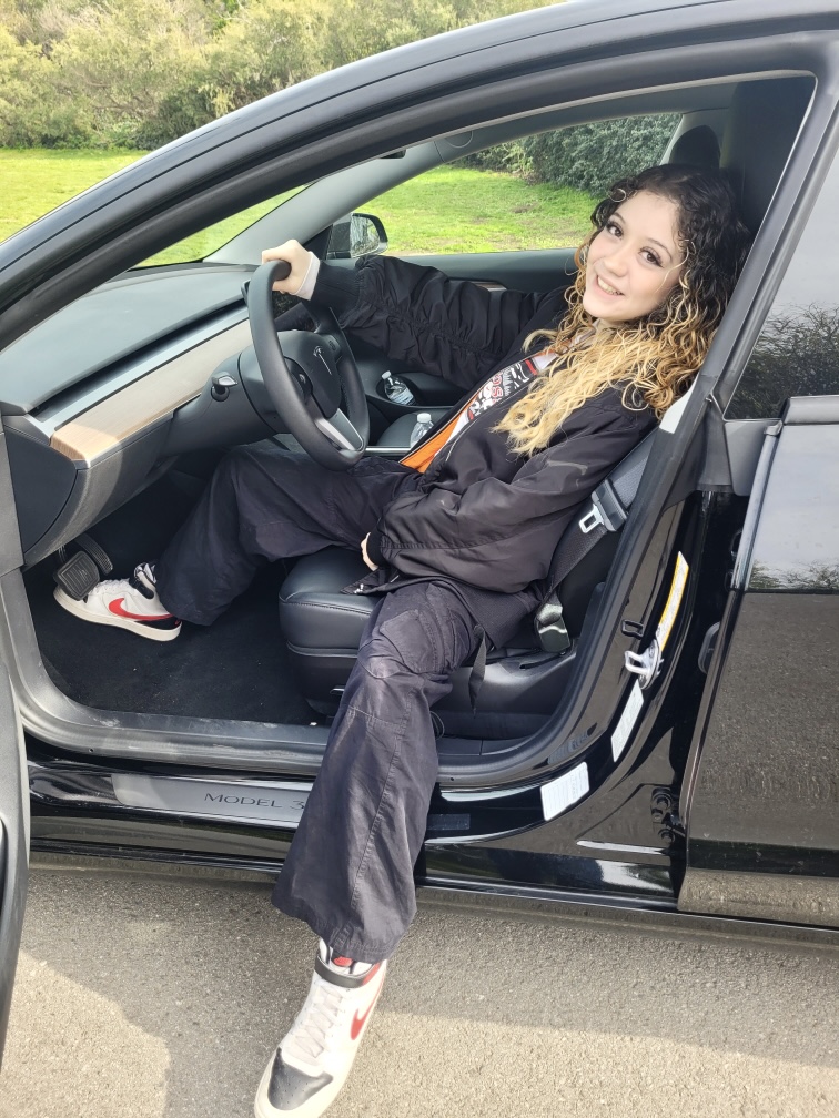 Emily Halasz sits in the driver seat of a parked Tesla, one leg outside the car.