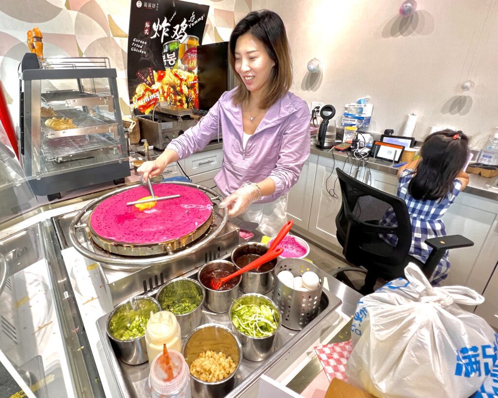 A woman prepares a Korean-style dragon fruit pancake with a tool specializing in flattening and spreading pancake batter as her daughter plays behind her.