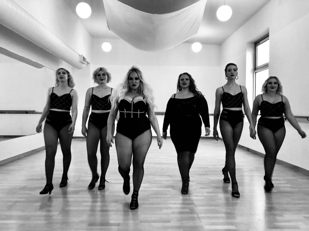 A black and white photo of the group Sugar Rush strutting down a dance studio practice room all dressed in black clothes and fishnet stockings.