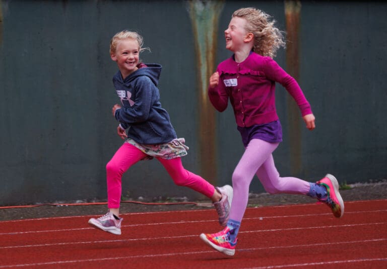 Annika Hardesty, 6, left, and Emma Parine, 5, laugh as they run on the red track field wearing different shades of pink and a dark blue jacket.