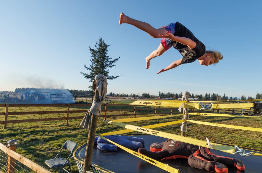 Rubin Hutchins, upside down midair, performs a Swanton Bomb wrestling move as firefighters put out a shed fire in the background.