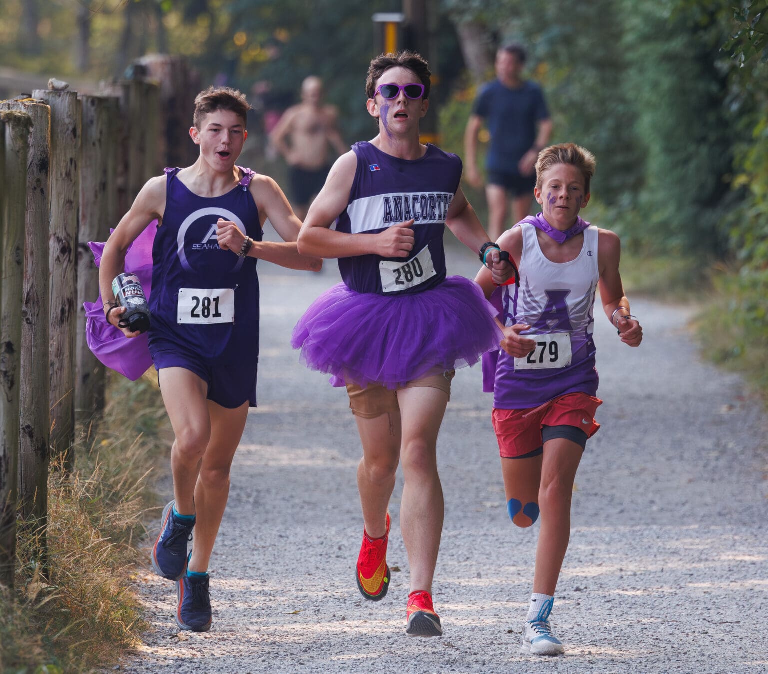 Maverick Ball, left, and Owen Kennedy, right, run with teammate Owen Hofheimer as he finishes the final lap dressed in their signature purple outfits, one of which is a tutu.