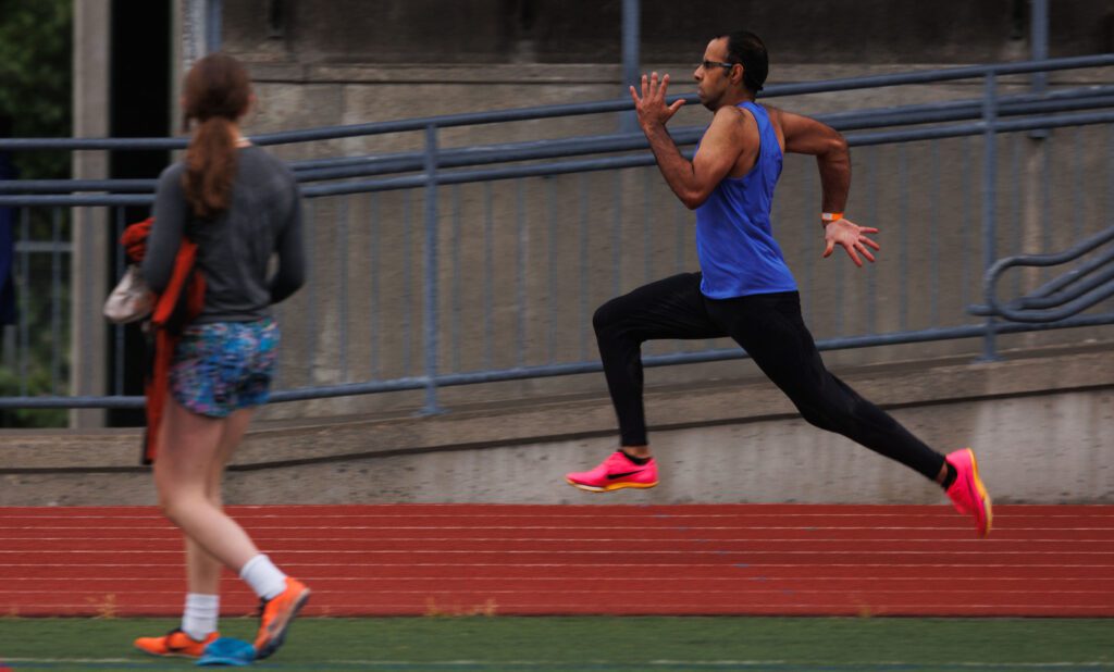Sunny Gill, 31, sprints across the red track field in pink sneakers, blue tank top and black pants as a woman watches him from the sidelines.