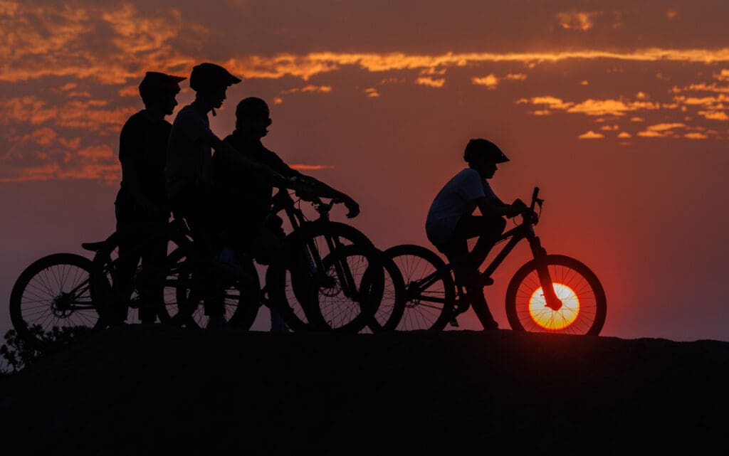 A red sun sets inside a bike rim as pump track riders wait their turn to ride with the dark orange sky behind them.