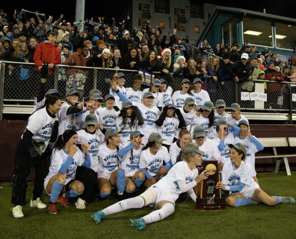 The Western Washington University women's soccer team poses with the national championship trophy with fans cheering and smiling behind them from the bleachers.