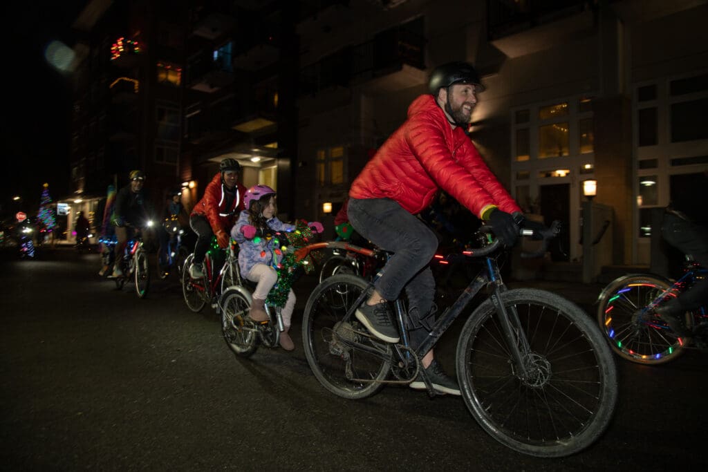 With lights and holiday decorations, cyclists pass by cars on the road.
