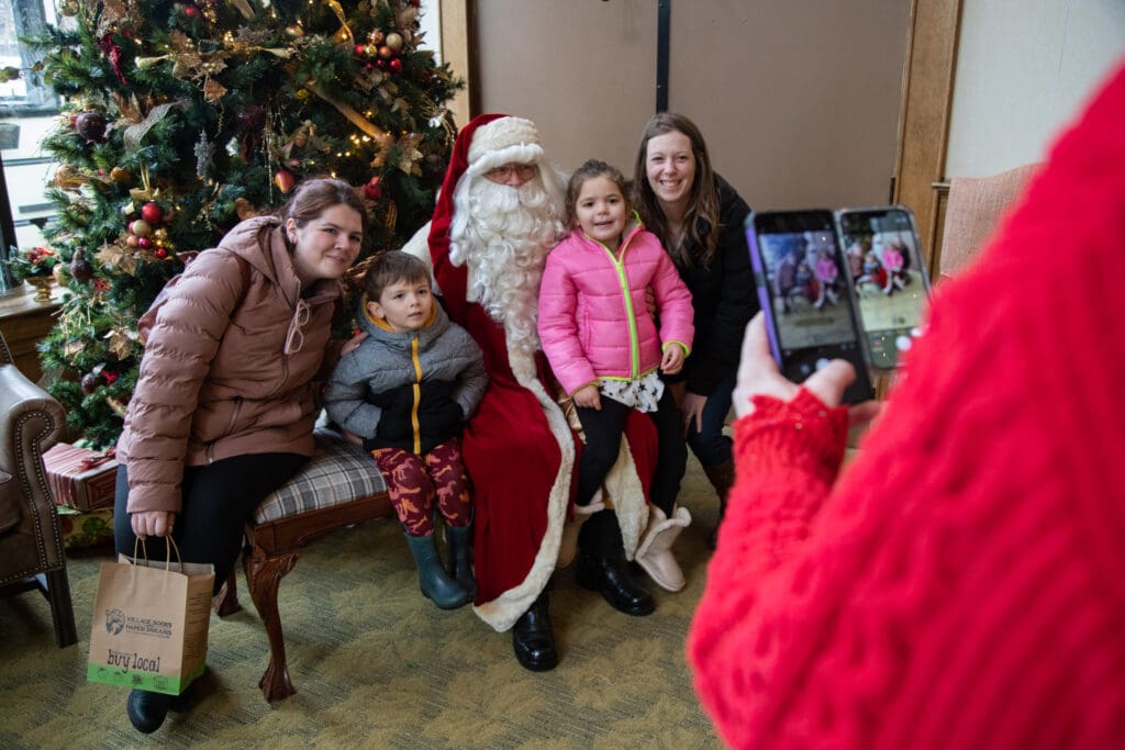 A person in a bright red sweater takes a picture for a family posing with Santa claus in front of the christmas tree.