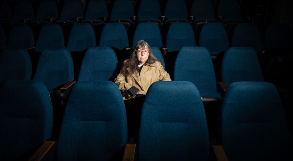 Susie Purves sitting alone in a theatre on one of the many rows of blue chairs.