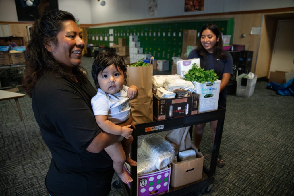 A mother holding her toddler next to a black tray stacked with boxes of necessities pushed by a woman.