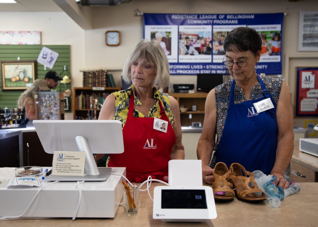 Two women in aprons stand behind a counter and ring up a pair of shoes on a point of sale system at a thrift shop.