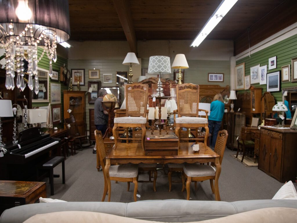 Old furniture — lamps, tables and chairs — are stacked up in a thrift store.