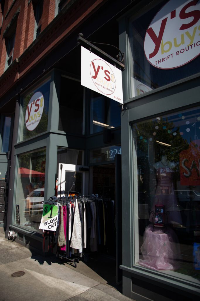 The exterior of a storefront dons a sign that reads "Y's buys thrift boutique." A rack of clothing stands outside the open door.