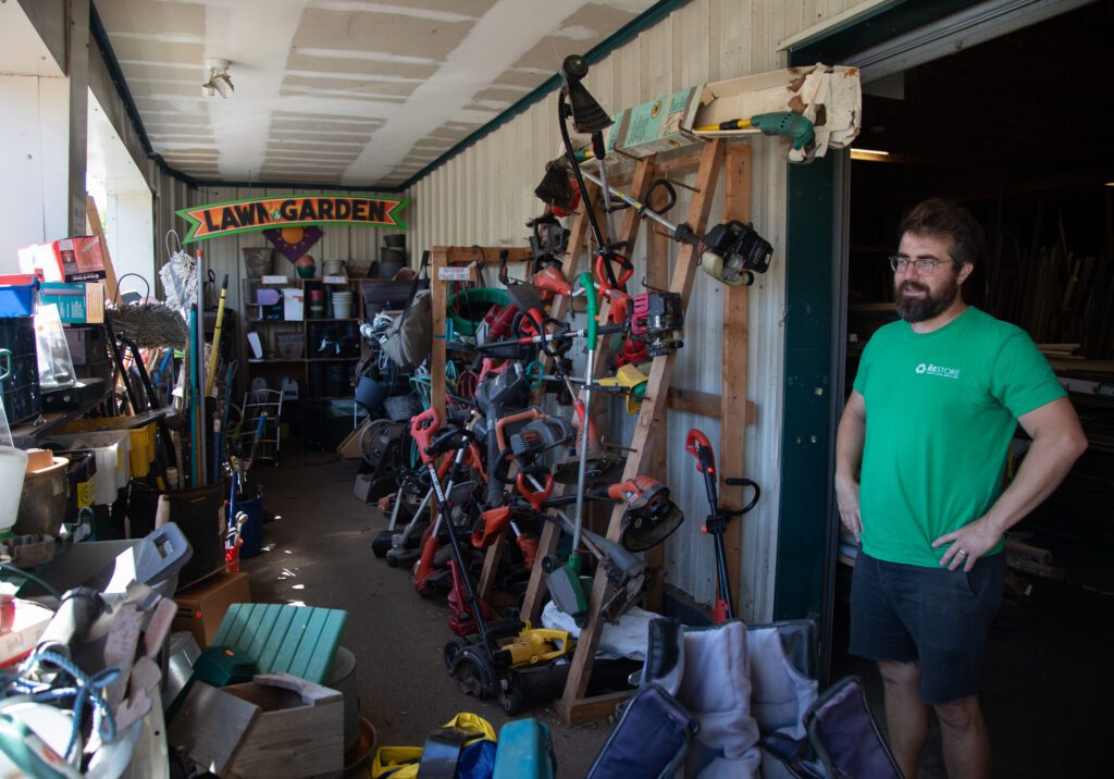 A bearded man in a green shirt and glasses stands with his hands on his hips in a room full of landscaping tools. On the wall is a sign that reads "Lawn & Garden."