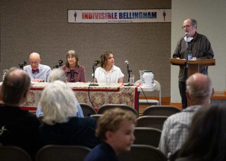 From left, former Bellingham School Board president Kenneth Gass, former Ferndale School Board president Lee Anne Riddle, current Bellingham director Jenn Mason and Indivisible Bellingham host Michael Berres sit on at a shared table facing the audience as guest speaker talks behind a podium.