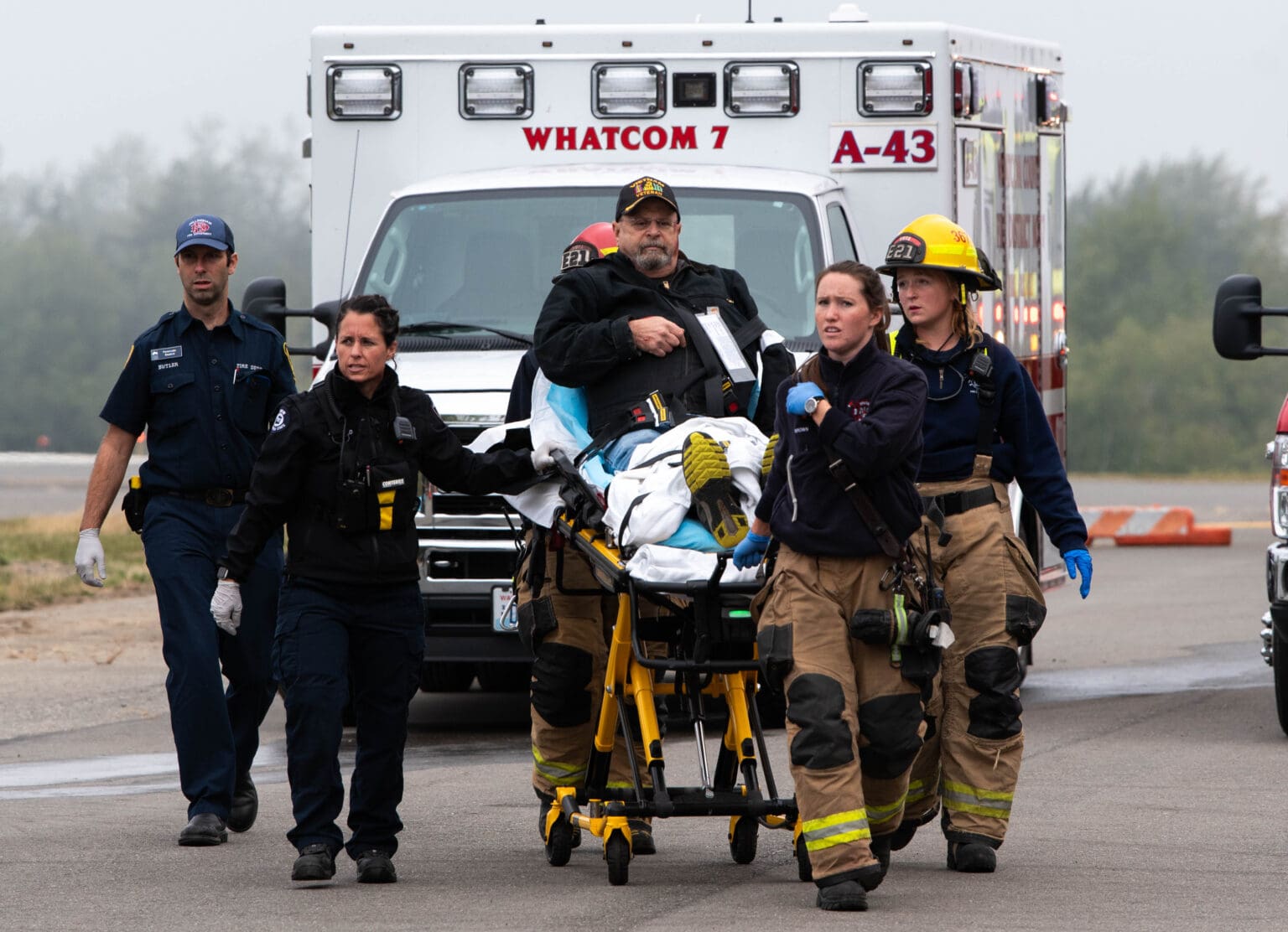 A volunteer victim on a stretcher gets escorted by multiple firemen in training away from the ambulance.