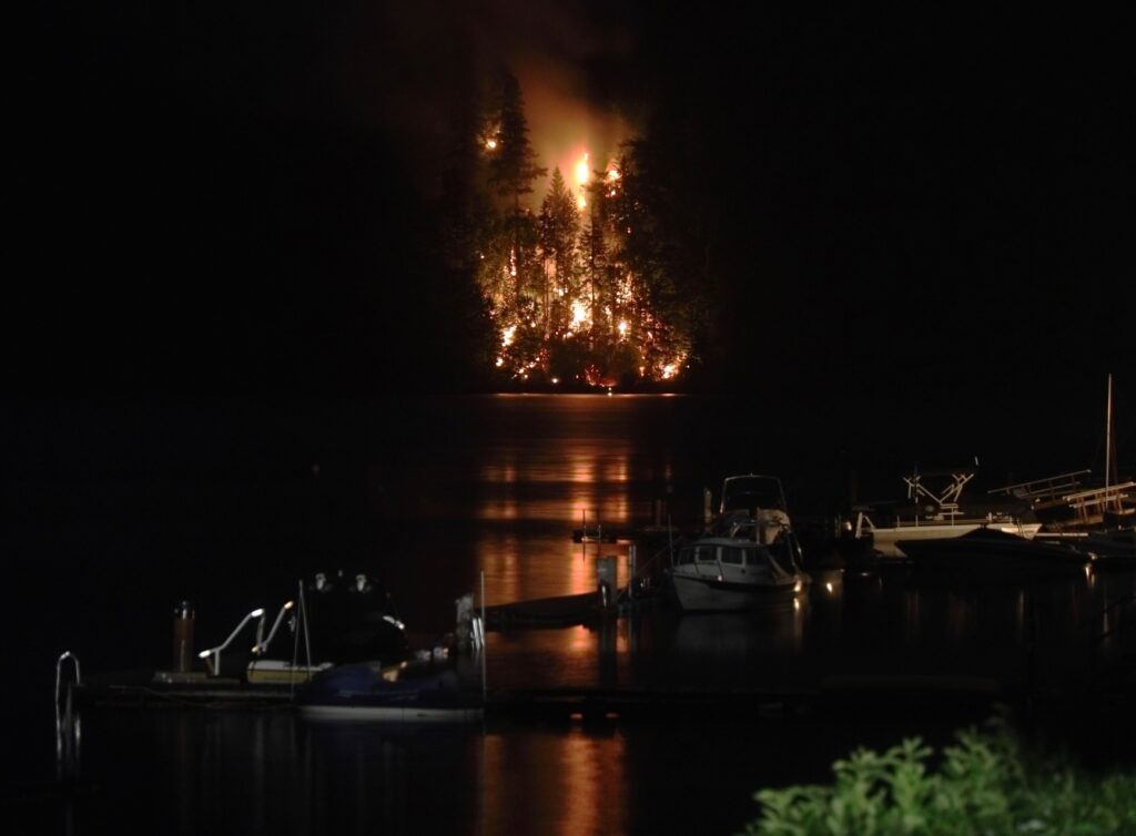A small wildfire burns a steep forest southeast of the shore of Lake Whatcom as boats are idle on the docks nearby.