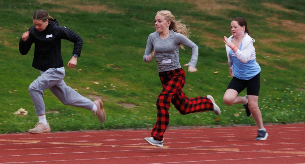 Three women spriint across the red track as they compete in sweats and jackets.