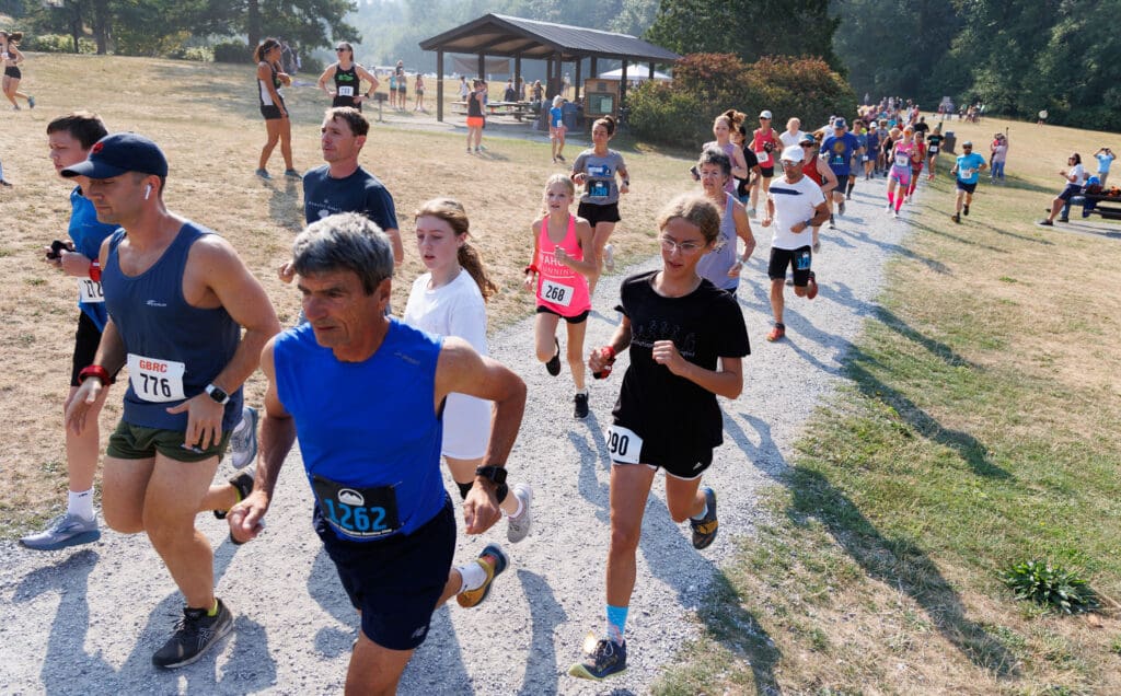 A large crowd of various ages and genders jog along a gravel path as spectators cheer them on from the sidelines.