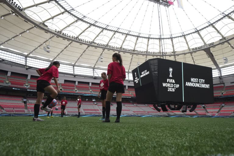 Soccer players in red shirts and black shorts pass soccer balls to each other in an indoor field which says "FIFA World Cup 2026, Host city announcement."