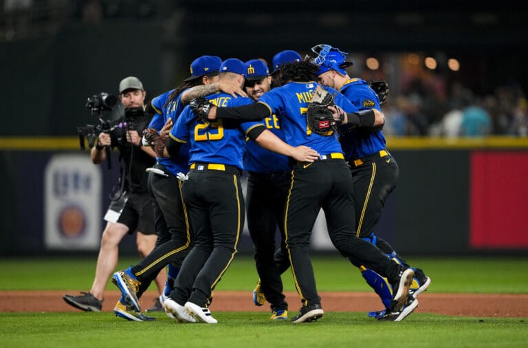 Seattle Mariners, including first baseman Ty France (23), third baseman Eugenio Suárez, center facing, and relief pitcher Andres Munoz (75) hold each other by the shoulder as they dance in a circle.