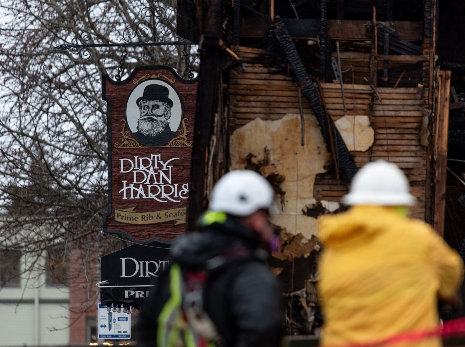 The sign for Dirty Dan Harris Steakhouse hangs in the background as a pair of construction workers inspect the burned and demolished parts of the building.