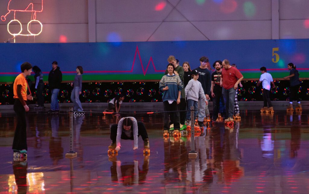 Roller skaters line up for a game of limbo as a participant attempts by squatting close to the ground.