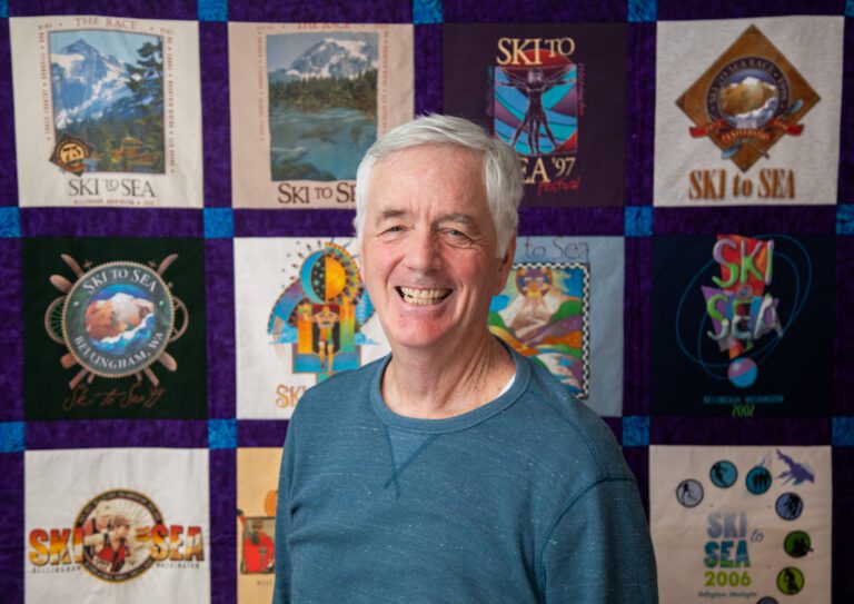 Longtime volunteer John Burley stands in front of a quilt of his Ski to Sea shirts.