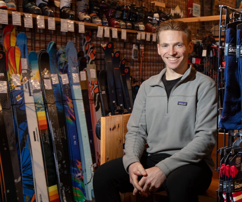 Luke Sutton sitting next to rows of different colored skis inside the ski shop.