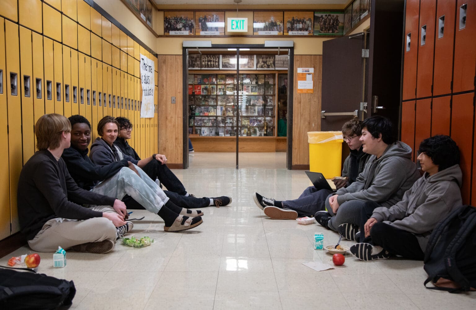 A group of students eat their lunch in the hallway outside the cafeteria with their backs to the lockers.