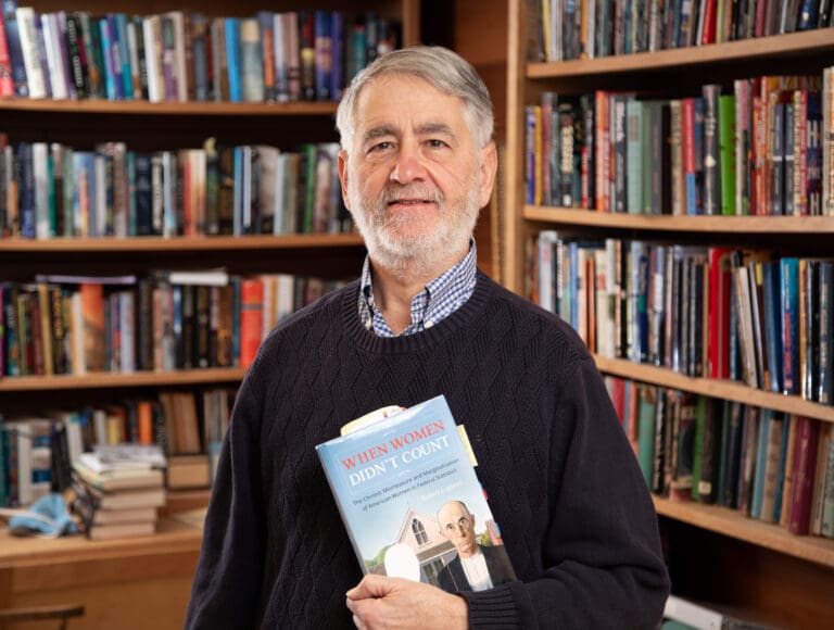 Author and retired librarian Rob Lopresti poses for a photo with a book in his arms as he is surrounded by shelves filled with books.