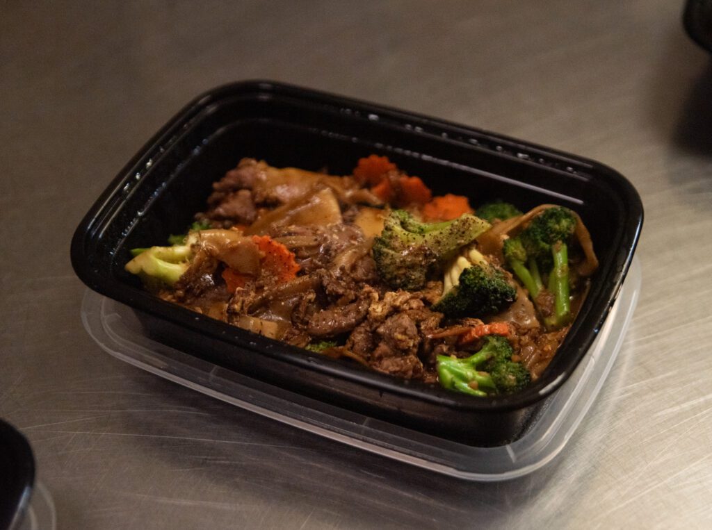 A takeout container of phad see ew, a dish served with stir fried vegetables, meats and flat noodles.
