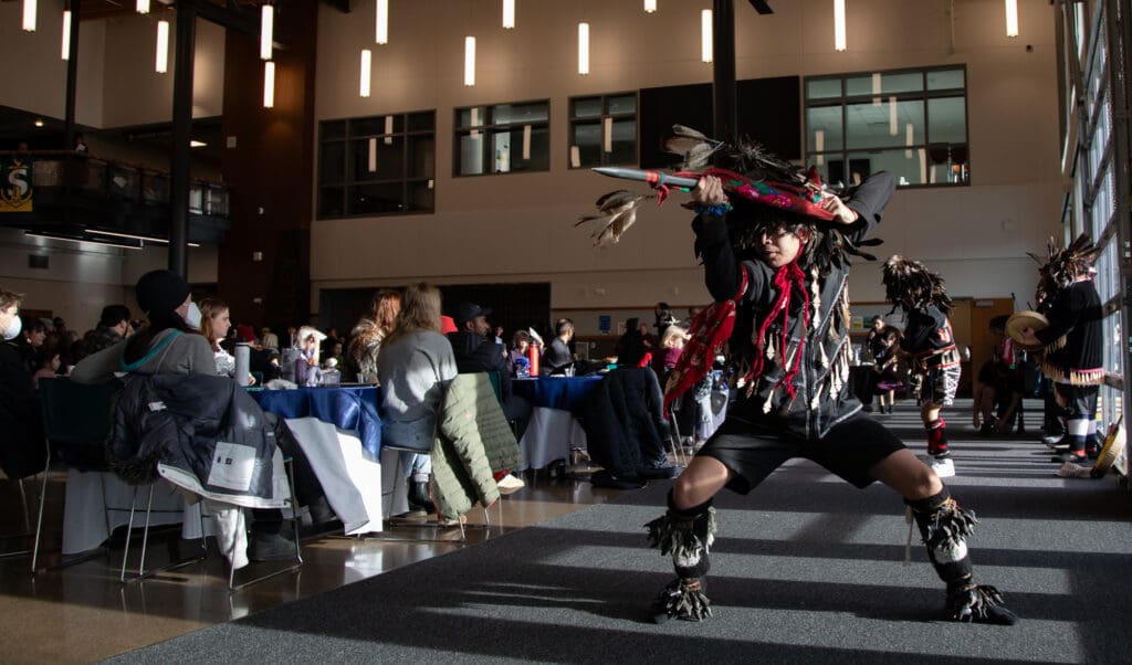 The Lummi Nation Blackhawk Dancers sing and dance at the MLK Day event breakfast wearing traditional clothing in front of their band of musicians.