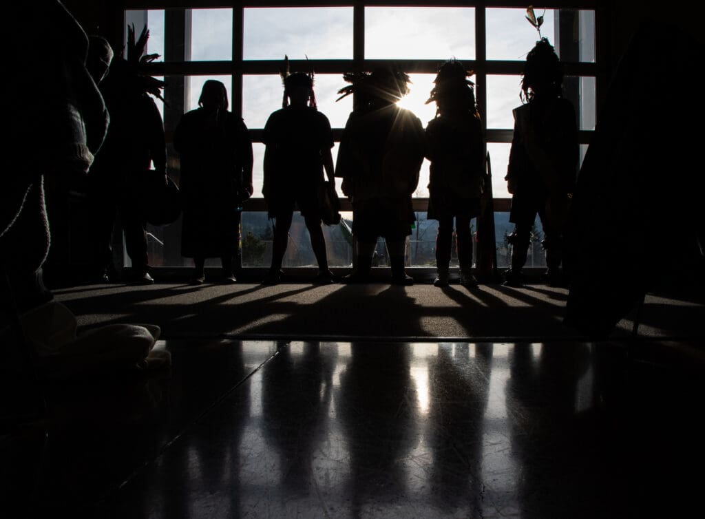 The Blackhawk Dancers stand in front of the windows letting the sun hide them only to show their silhouettes.