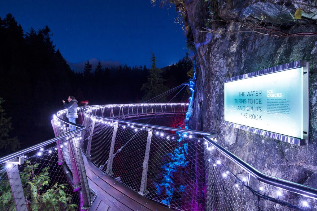 A person takes a photo on the brightly lit railing connected to the cliff behind him.