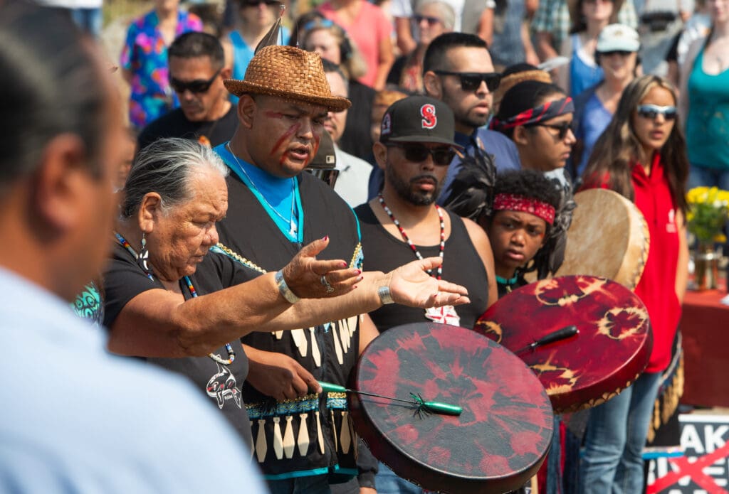Members of the Lummi Nation sing to honor Tokitae (also know as Sk’aliCh’elh’tenaut) as some play traditional drums while a crowd watches.