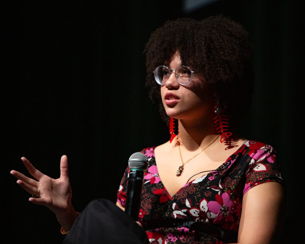 Izabella McFrazier gestures as she talks into a microphone dressed in red pink floral clothing and fish bone earrings.