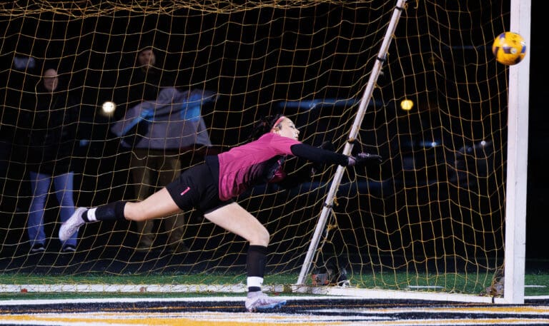 Blaine goalie Larissa Pluschakov leans hard to block the ball from scoring a goal as spectators from behind the net watch.