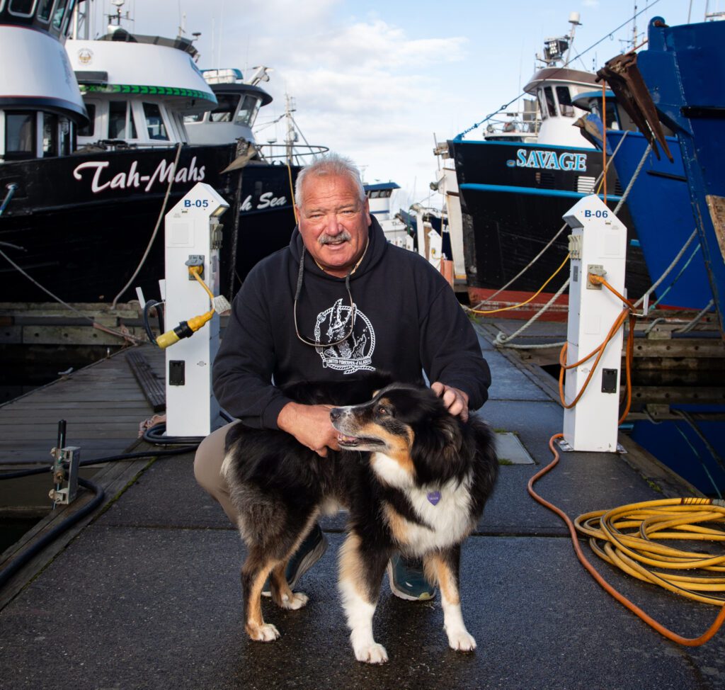 A man petting his dog on the docks inbetween docked ships.