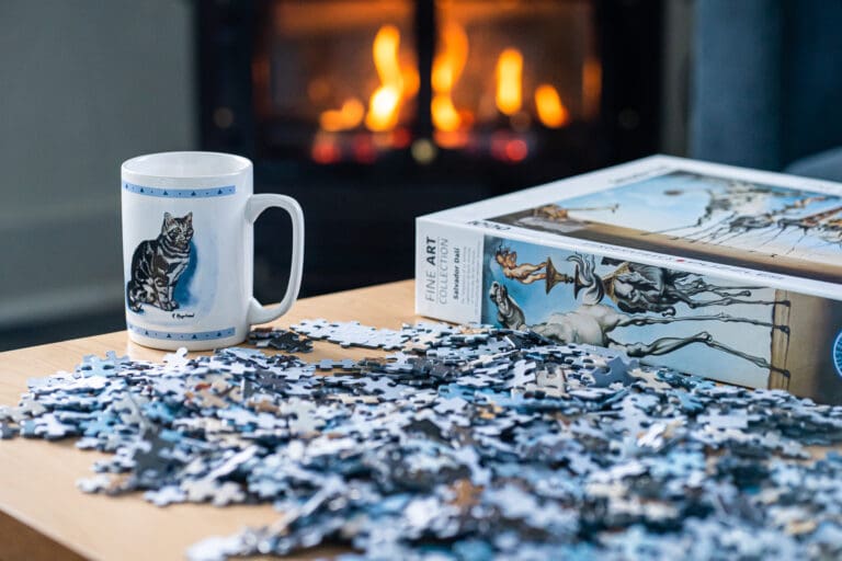 A mug with a cat printed on sits next to an unfinished puzzle as a fireplace behind the items warms up the room.