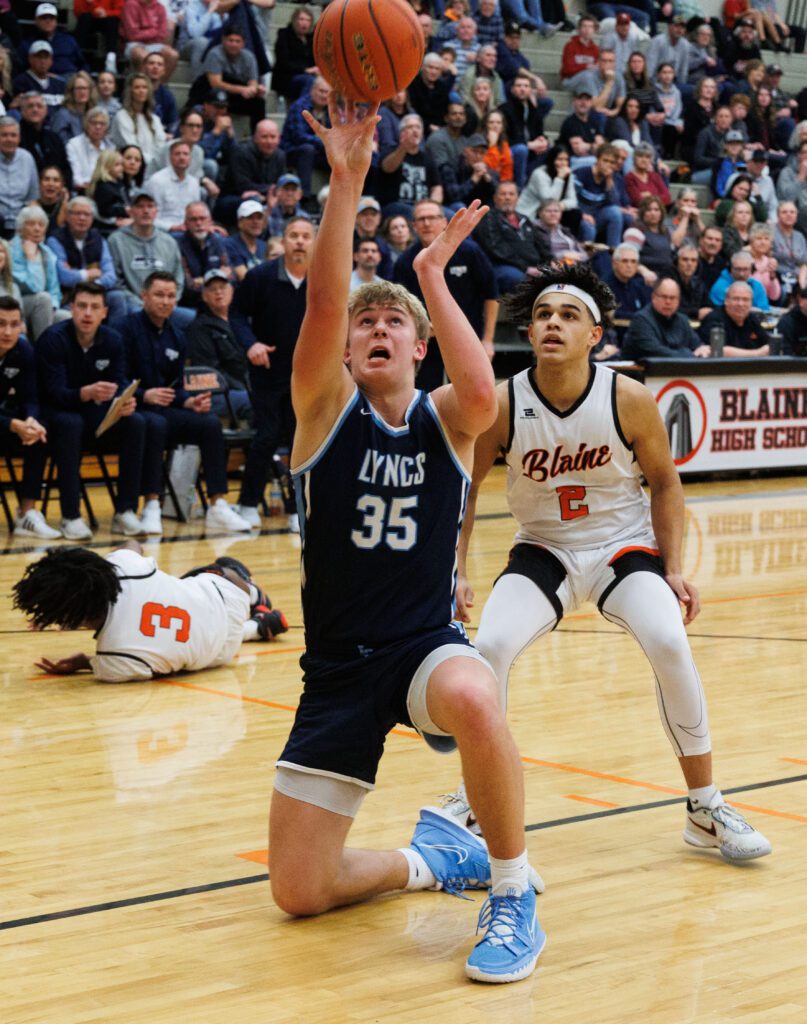 Lynden Christian's Jeremiah Wright attempts to make a basket while on his knee as a defender looks up to the ball while another player is left on the floor.