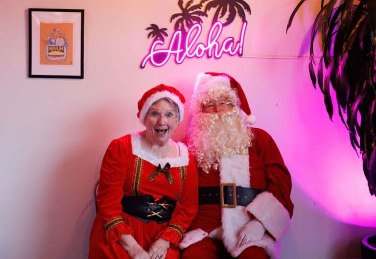 Lynn, left, and Doug Starcher pose as Mrs. Claus and Santa Claus underneath a neon "Aloha" sign.