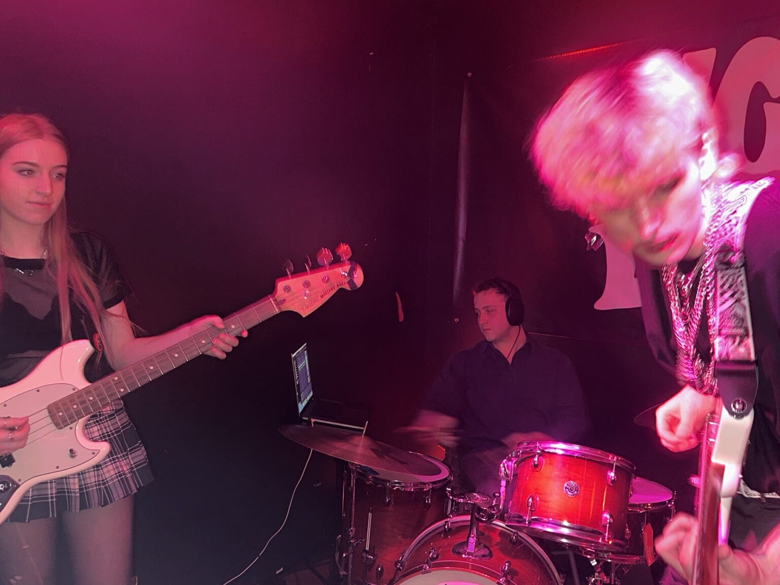 Band members of Girls Knows playing their instruments under a bright pink light.