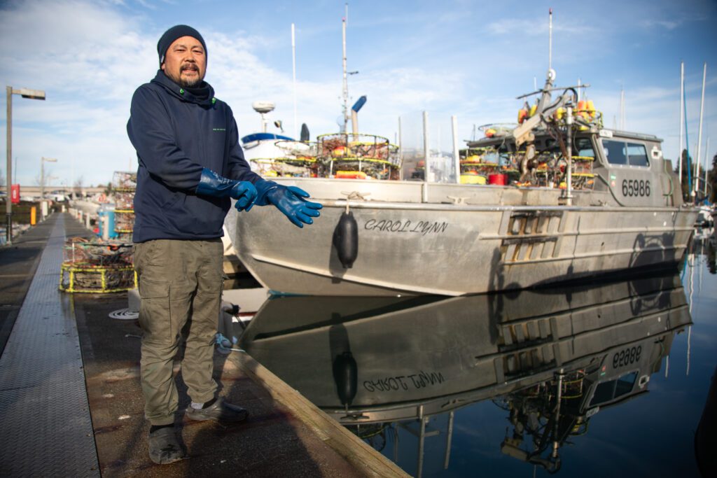 A fisherman stands near a docked boat while he removes his rubber gloves.