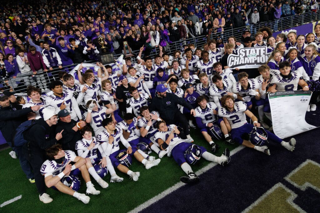 Anacortes players pose for a photo in front of students and fans while holding up trophies and a sign declaring them State Champions.
