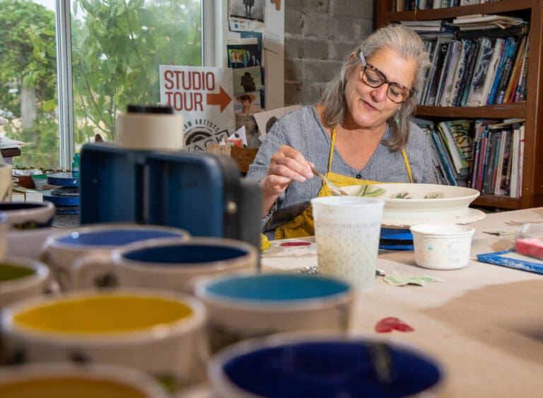 Ann Marie DeCollibus glazes a plate in her home studio surrounded by multiple art supplies and various paints.