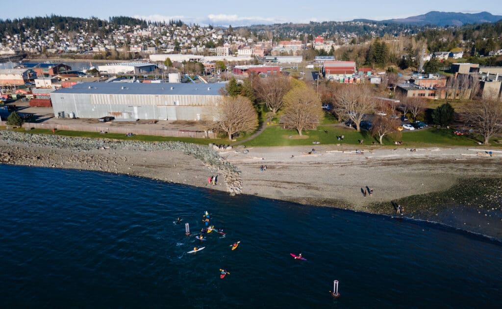 An aerial view of Kayakers playing water polo in Marine Park in Fairhaven with trees and the city of Bellingham in the background.