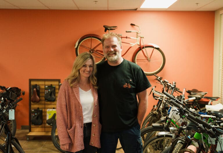 Bike enthusiasts John and Carolyn Roy inside their Birch Bay Bike Shop with a bike mounted on the orange wall behind them.