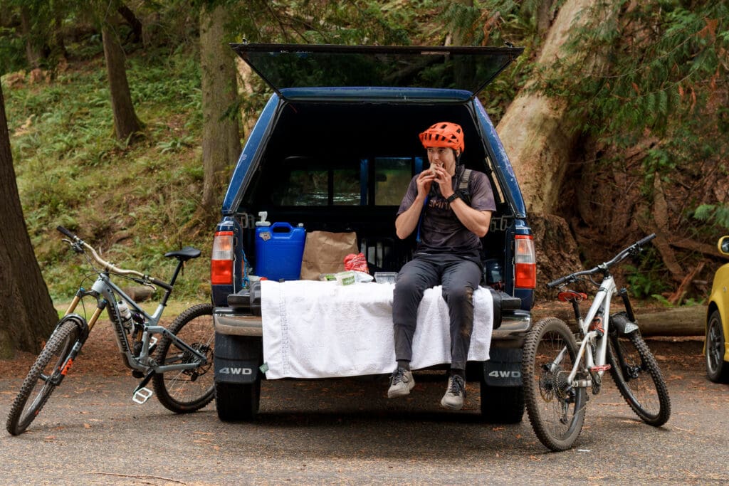 Talus Lantz wearing his orange cycling helmet sits on the back of his car as he eats to refuel.